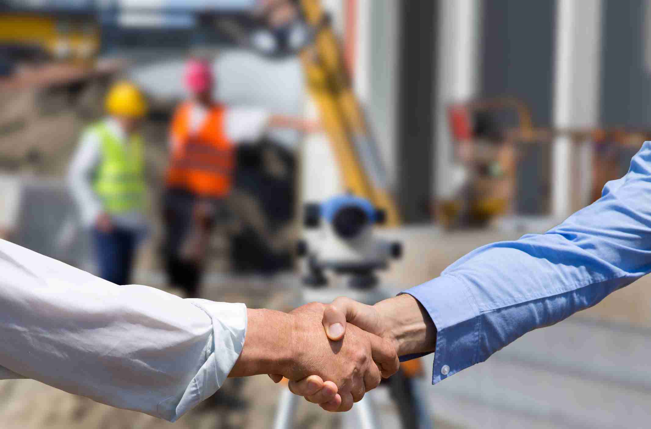 Two business people shaking hands in front of leveling instrument on construction site with workers in background