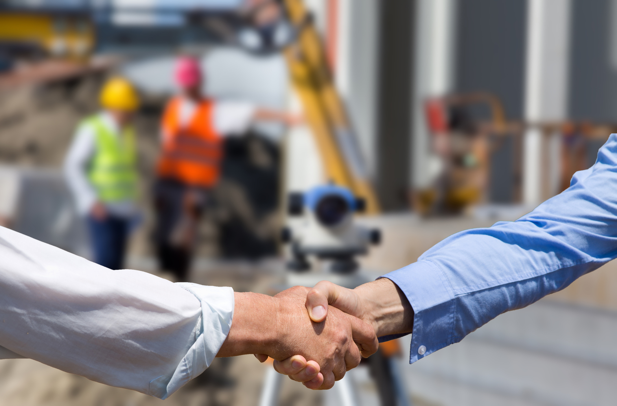 Two business people shaking hands in front of leveling instrument on construction site with workers in background