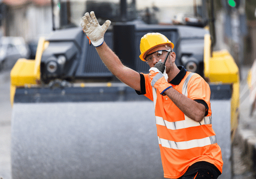 A construction worker in a bright safety vest and hard hat is signaling for vehicles to stop while overseeing roadwork in a busy urban area.