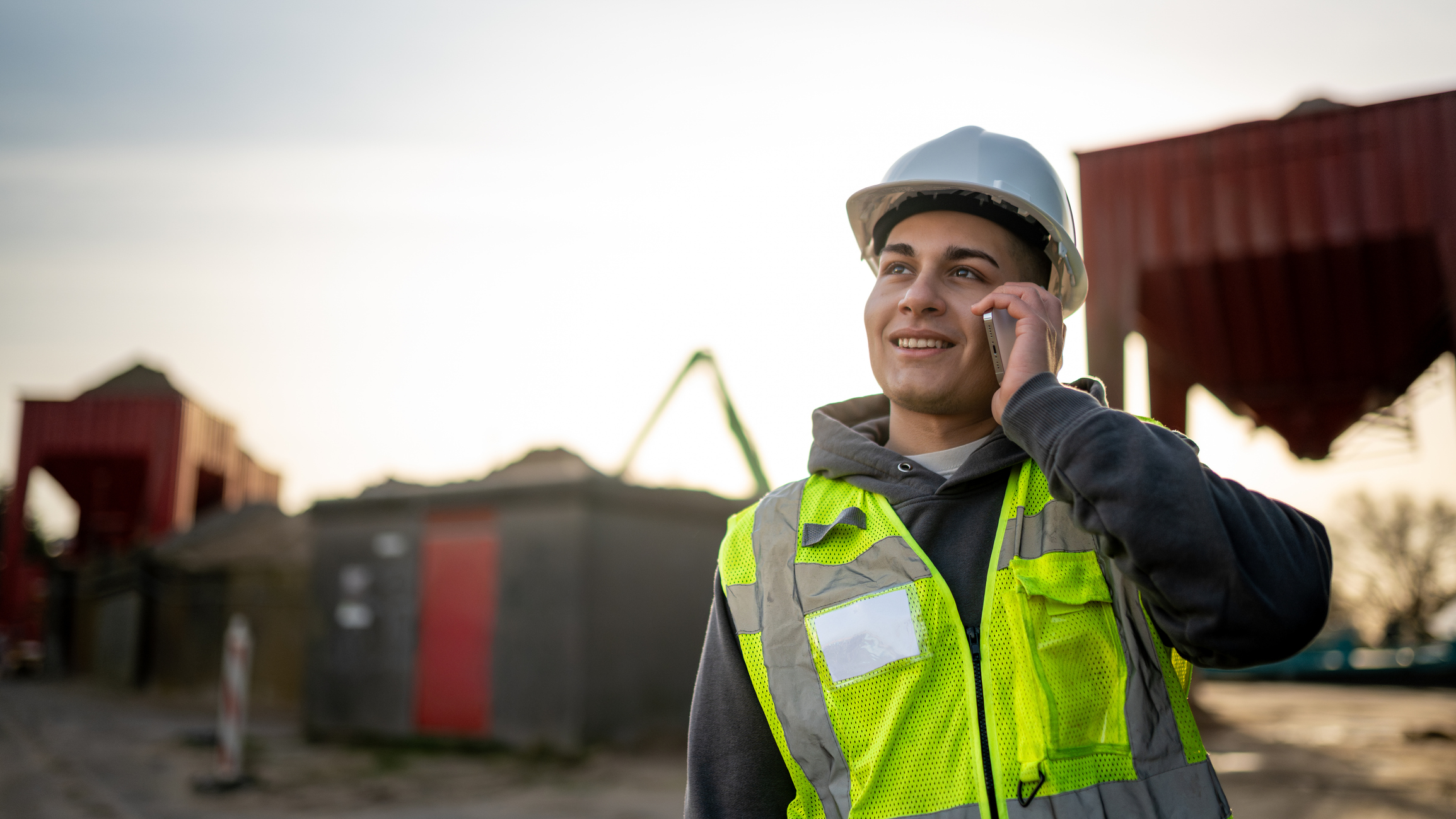 Junior engineer having a phone conversation while working on a construction site, ensuring safety with his hard hat and reflective vest